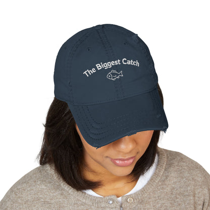 Navy blue cap with 'The Biggest Catch' text worn by a person on a white background